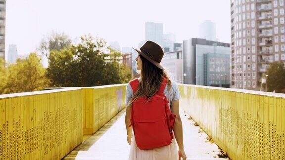 戴着帽子长发的女孩走在城市的桥上一个背着红色背包站在黄色墙壁中间的白人女人后视图4k