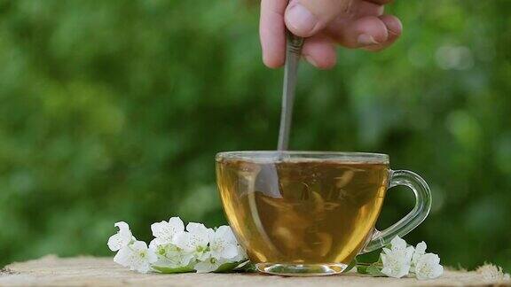 一杯茉莉花绿茶茉莉花茶和茉莉花的木制背景茉莉花和叶子慢动作将茶倒入杯中用茶匙搅拌近距离