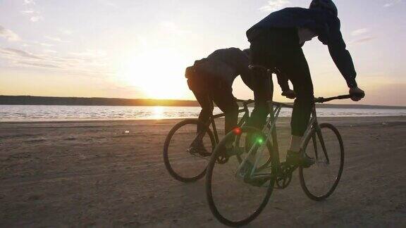 两个年轻人在海滩上骑着自行车背景是橙色的夕阳慢镜头
