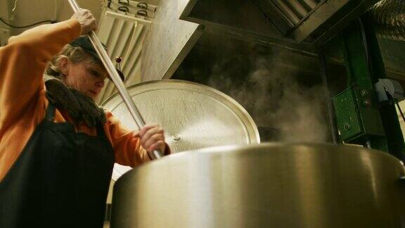 一名60多岁的白人妇女在一个商业厨房制造设施中用长桨在一个大型金属储存罐中搅拌液体食物