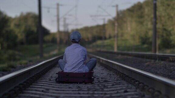 戴着帽子的伤心男孩坐在外面铁路上的一个手提箱上