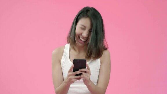 年轻的亚洲女性用智能手机查看社交媒体在粉色背景下穿着休闲服装感觉快乐的微笑快乐的微笑可爱的快乐的女人庆祝成功