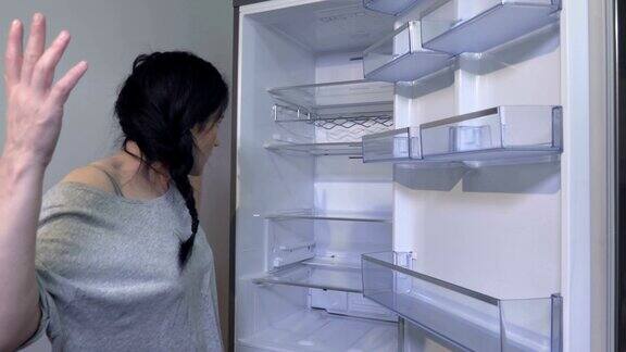 不快乐和饥饿的黑发女人站在冰箱旁边空的架子