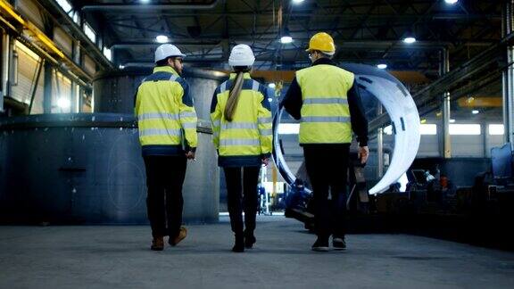 以下是三名工程师走过重工业制造厂的照片背景中各种金属制品管道桶组件缓慢的运动