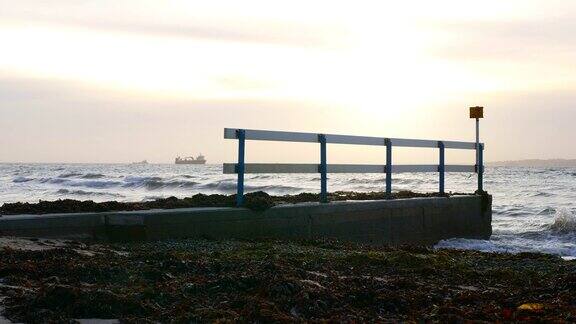 海浪拍打着码头和海草远处的货船在夕阳的背景下