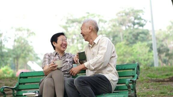 慢镜头快乐的亚洲老夫妇坐在早上的公园喝茶喝咖啡交谈并表达爱意