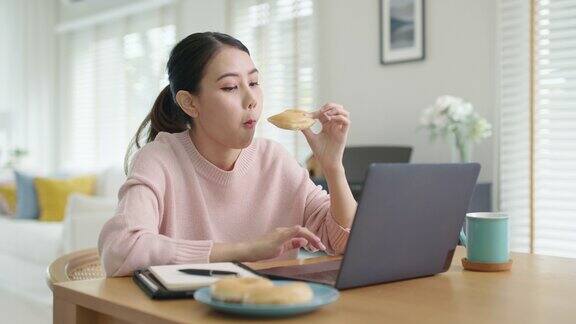 年轻迷人美丽的亚洲女性饿了吃甜甜圈吃零食吃满嘴看电脑笔记本在家里在忙碌的工作从家里多任务不健康的饮食生活理念