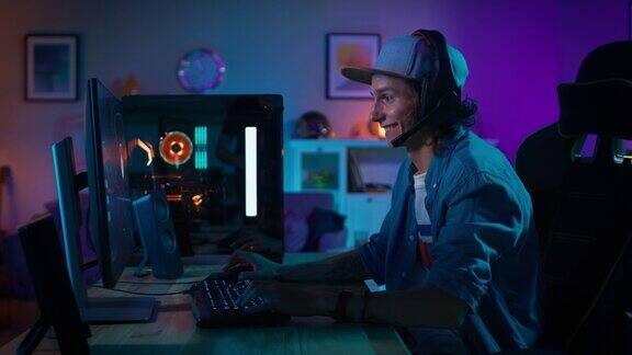 玩家戴上麦克风耳机开始在个人电脑上玩射击在线视频游戏房间和电脑都有彩色霓虹灯年轻人戴着一顶帽子在家度过一个舒适的夜晚