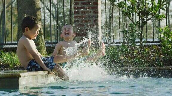 在一个阳光明媚的日子里两个白人小男孩(一个三岁一个五岁)坐在游泳池边一边在水里踢脚一边微笑和大笑身后实时有一个金属栅栏