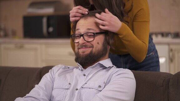 一个戴眼镜的大胡子男人正坐在沙发上一个黑发女人站在他身后把她的一缕头发放在他的前额当作刘海拥抱他