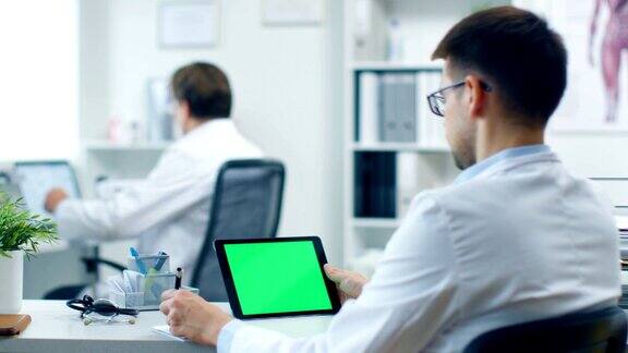 特写的一个男医生使用平板电脑与绿色屏幕他也拿着笔在他的记事本他的助手工作在背景办公室是明亮白色和现代