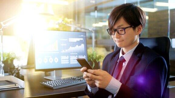 在办公室东亚商人使用智能手机输入重要信息他的台式电脑显示了公司增长的数据阳光普照