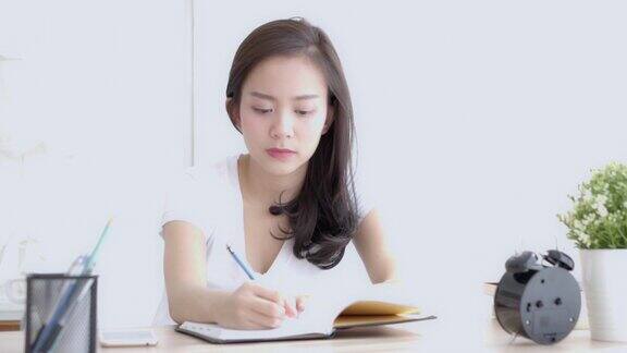 美丽的年轻亚洲女人微笑着坐在客厅学习和学习写笔记本和日记在家里女孩家庭作业女商人计划工作在桌子上教育理念
