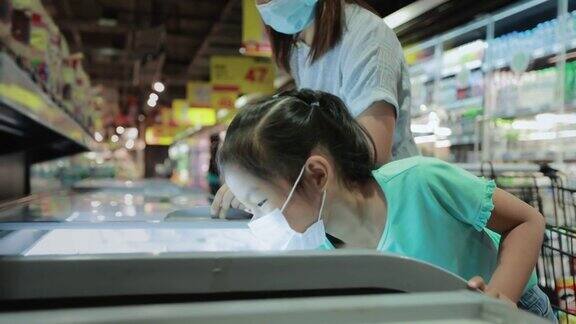 一名亚洲女童和她的母亲戴着防护口罩以预防冠状病毒大流行新的正常要求是一起选择超市冷藏室的冷冻食品