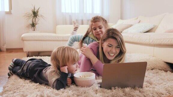 快乐的妈妈和她的两个孩子使用笔记本电脑在客厅吃爆米花