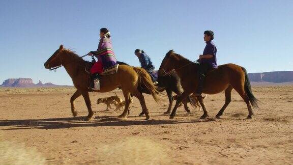 几个年轻的印第安人(纳瓦霍)儿童骑着马通过纪念碑谷沙漠与他们的宠物狗在亚利桑那州犹他州在一个晴朗明亮的日子