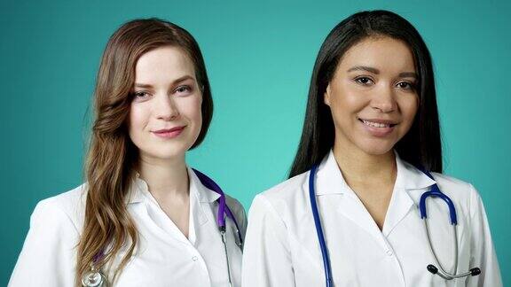美丽的年轻快乐的女孩对着镜头微笑两个成功自信的现代女医生穿着白大褂摆姿势慢动作女性医学工作者保健和医学概念