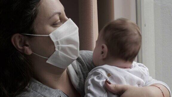 母亲戴着防护口罩抱着她的小婴儿她透过窗户看