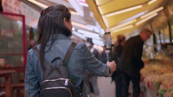 亚洲女性博主在城市的露天市场用自拍杆拿着手机录制视频年轻的亚洲女游客女子手持智能手机录制视频旅游概念