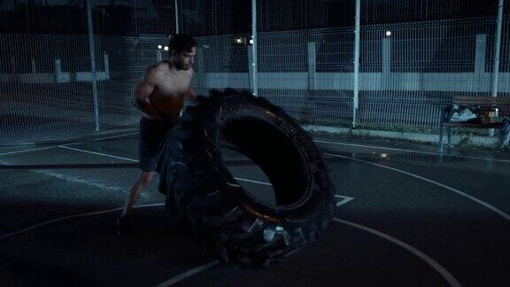 强壮的肌肉健康年轻的赤膊男子正在做运动在一个围栏室外篮球场在一个雨后的夜晚他在居民区掀翻一个沉重的大轮胎