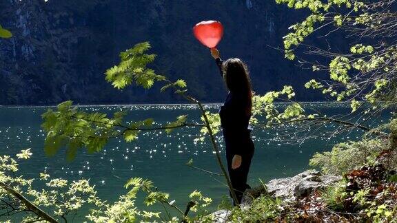 小女孩拿着心形的红色气球站在湖边