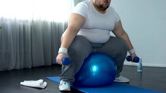 肥胖男性穿着运动服坐在健身球上举哑铃活动