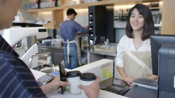 在咖啡馆里一位美丽的亚洲女人正在为一位通过非接触式手机向信用卡系统付款的顾客制作外卖咖啡