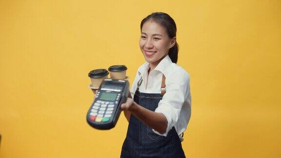 快乐微笑的亚洲女服务员穿着围裙友好地看着相机给顾客咖啡或茶和信用卡读卡机孤立在黄色背景