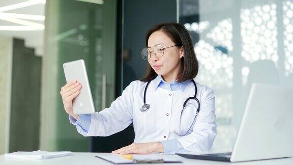 在现代医院的诊所里穿着白大褂的亚洲女医生正在用平板电脑进行视频通话
