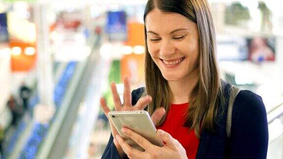 年轻漂亮的女人站在购物中心微笑用她的智能手机和朋友聊天