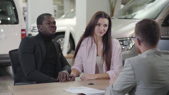 年轻美丽的白人妇女与汽车经销商交谈并握着丈夫的手混血儿家庭购买新车一对快乐的混血儿在陈列室做交易汽车经销汽车生意
