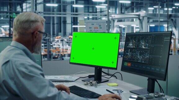 汽车工厂监控办公室:男首席汽车工程师坐在办公桌前工作的绿色屏幕色度键电脑自动化机器人手臂装配线制造车辆