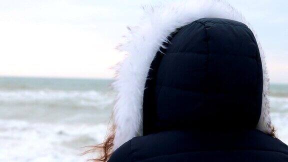 从后面看一个穿着冬季大衣带着兜帽的女人在看黎明前的海浪特写
