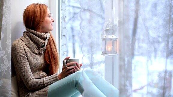 一个红头发的年轻女子坐在窗台上看着窗户喝着茶窗外是冬天
