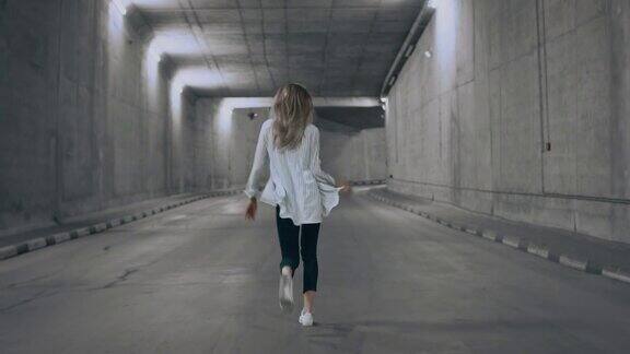 快乐美丽凉爽年轻的金发女人是走和跳舞在一个照明的混凝土隧道她穿着白色条纹衬衫和深色牛仔裤