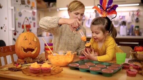 好奇的女儿正在观察她的母亲她正在用绳子上的橙色糖衣装饰一个万圣节纸杯蛋糕