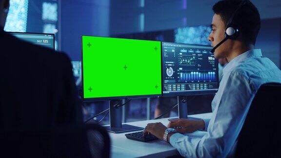 自信的男性数据科学家工作在个人电脑与绿屏模板戴着耳机在大控制和监控室呼叫中心办公室里的年轻工程师和同事们
