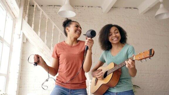 混合种族的年轻有趣的女孩跳舞与吹风机和弹吉他在床上唱歌姐妹们有乐趣休闲在卧室在家