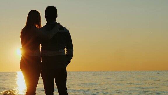 一对年轻的夫妇在海边拥抱着欣赏夕阳的剪影后视图