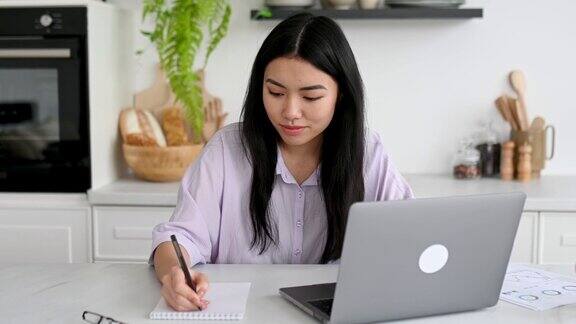 聪明可爱、积极乐观的亚洲女孩穿着衬衫设计师自由职业者或学生坐在家里的厨房里拿着笔记本电脑远程工作或学习做笔记分析友好的微笑