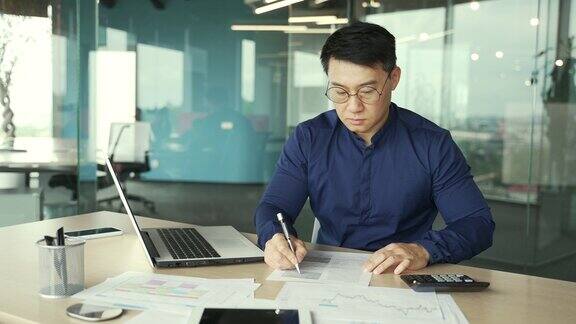 忙碌的亚洲会计文员在现代玻璃办公室的桌子上处理文件集中的簿记员用文件在计算器上计算财务报告