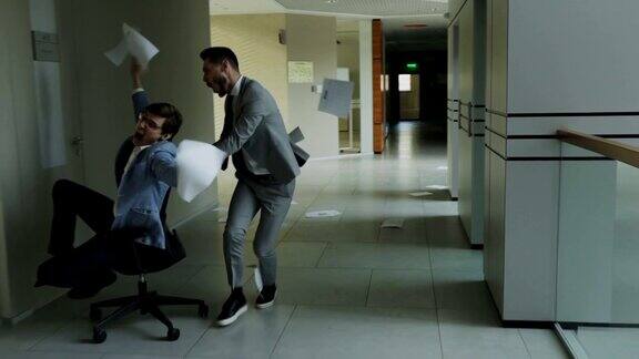 慢镜头拍摄了两个疯狂的商人坐在办公椅上在现代商业中心的大厅里玩得很开心把文件扔了起来