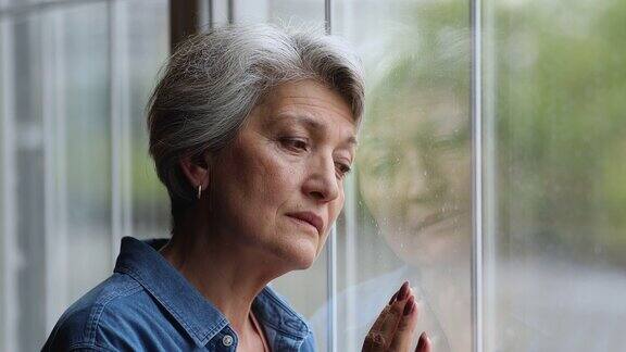 悲伤的老妇人站在窗边看着窗外感到很沮丧