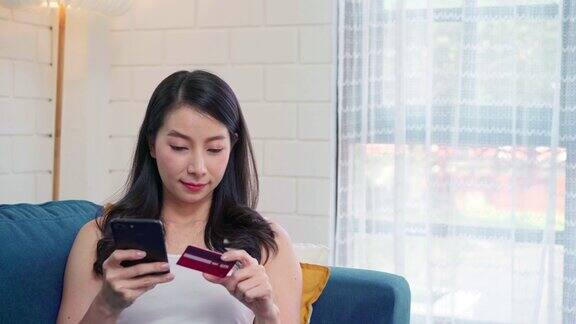 年轻微笑的亚洲女子使用智能手机在网上购物当躺在沙发上在家客厅放松时用信用卡购物居家女性的生活方式概念