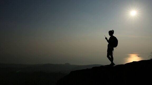 女徒步者用智能手机拍照在日落的山峰悬崖边缘