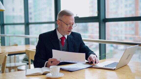 一个商人坐在桌子前检查打印出来的报告或文件用笔记本电脑解释他的观点办公室里的视频