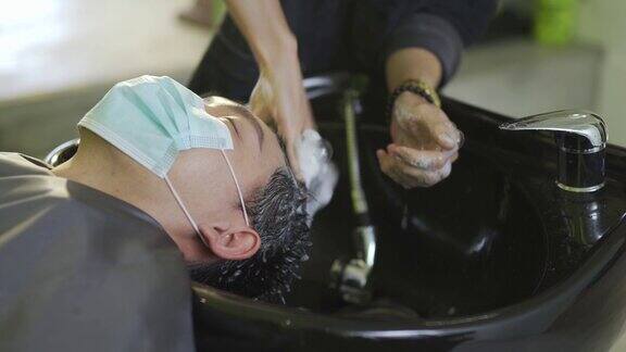 戴着保护面罩的亚裔华人男子在美发沙龙里闭着眼睛躺着洗头