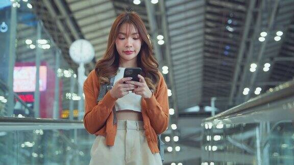 亚洲女游客背着背包站在机场候机楼的移动走道上使用手机旅游旅行