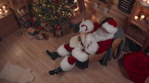 从高角度看圣诞老人在扶手椅上甜蜜地睡觉手里拿着长长的我们看到他在装饰华丽的房子里圣诞树上挂着彩灯
