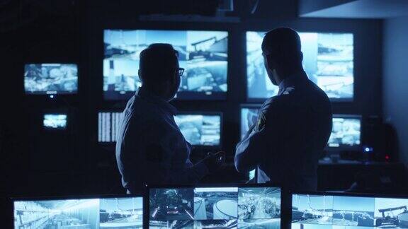 两名安全人员正在一间满是显示屏的黑暗监控室里交谈